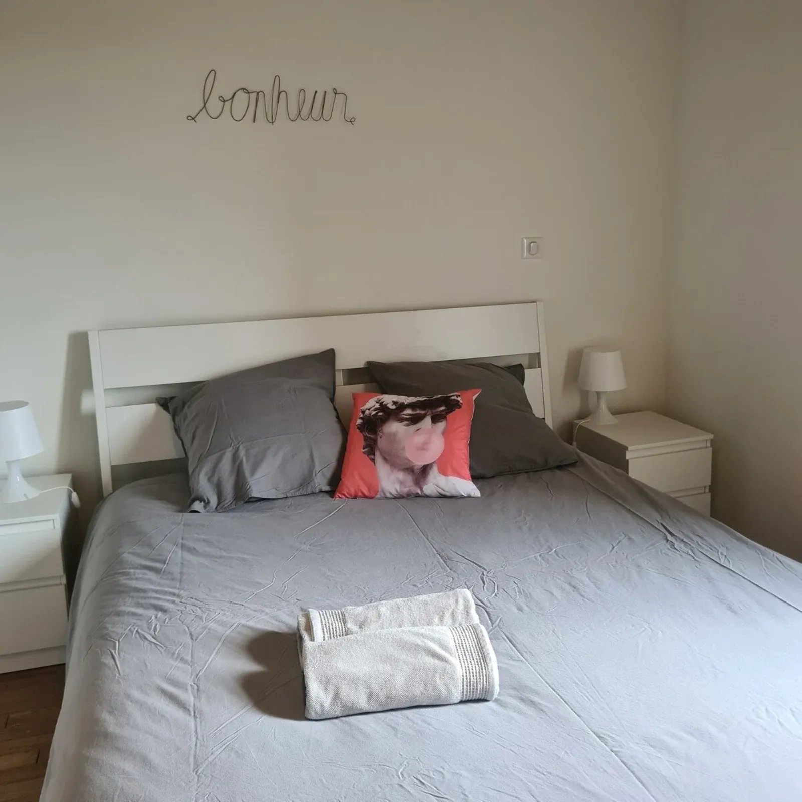 Bedroom with a double bed, grey linen and the word « bonheur » above the headboard