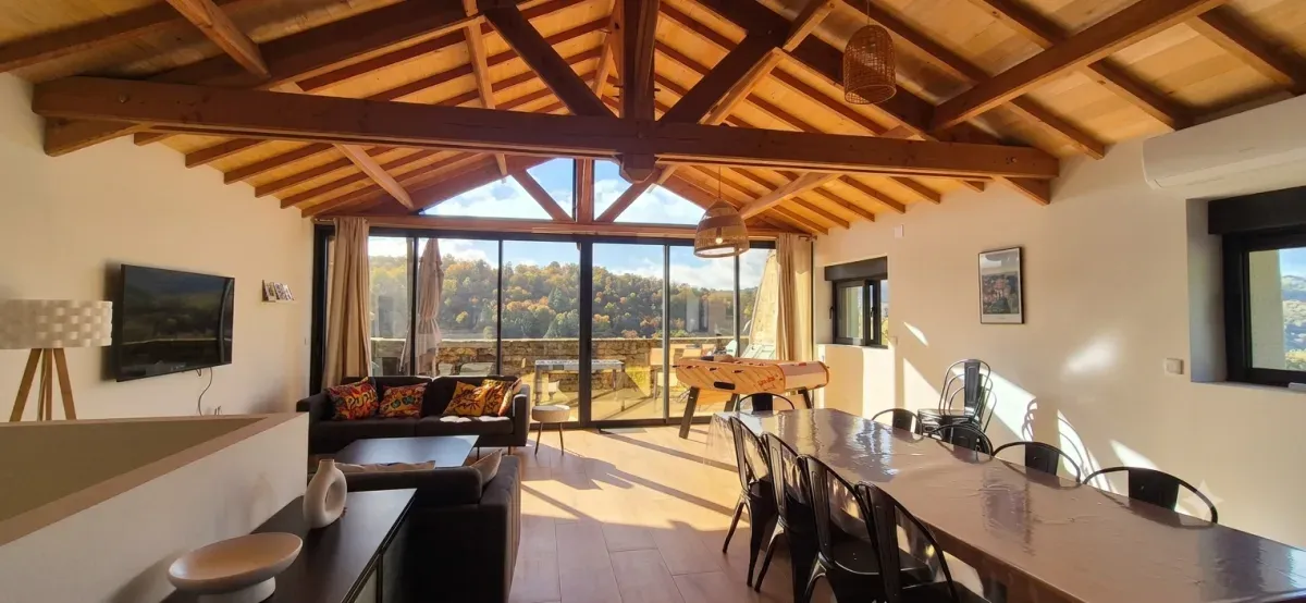 Large living room in the barn under exposed beams, with sofas, table football, a long table and picture windows onto the wooded valley