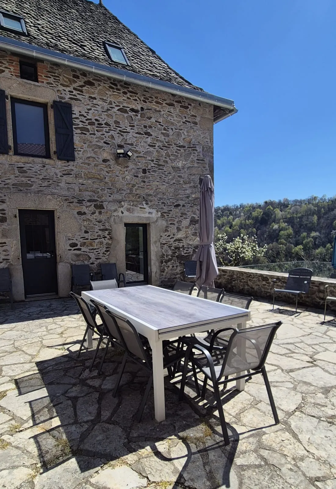 Paved terrace with a table for eight and a parasol in front of the stone house with grey shutters
