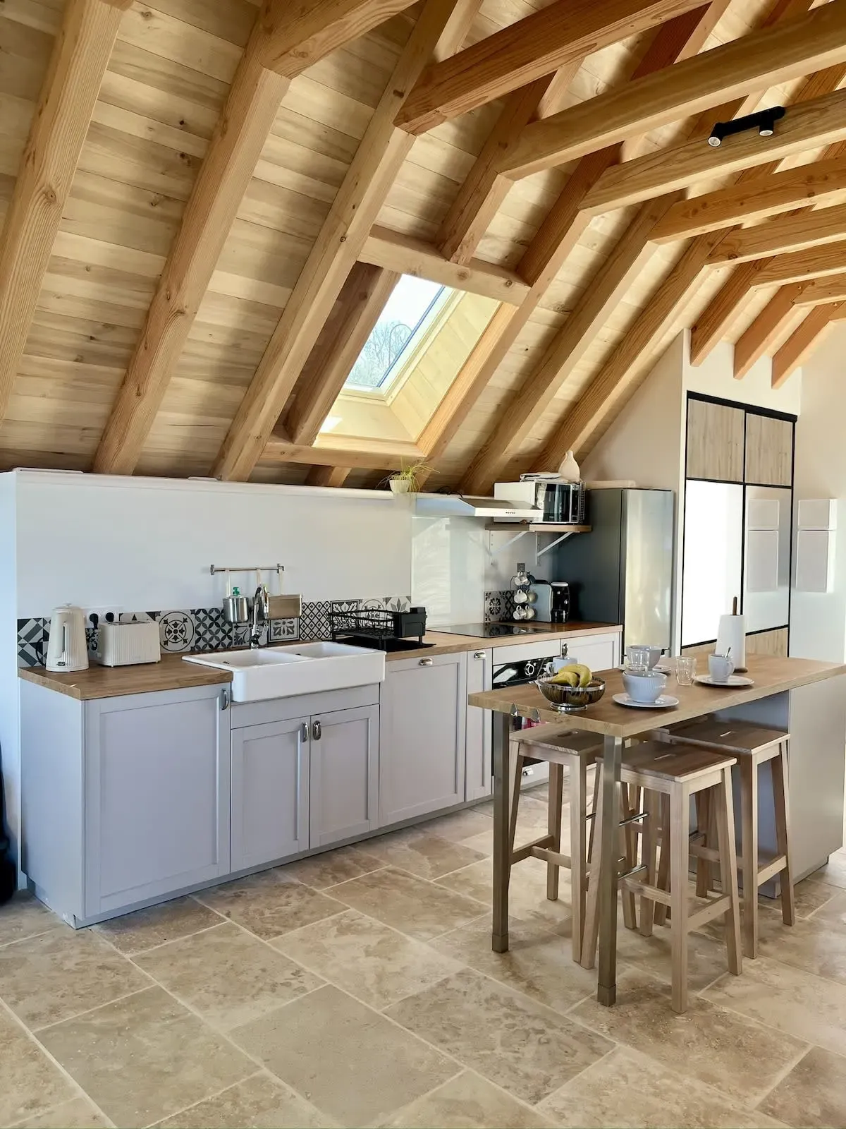 Cuisine équipée sous charpente, évier en céramique, îlot et tabourets en bois