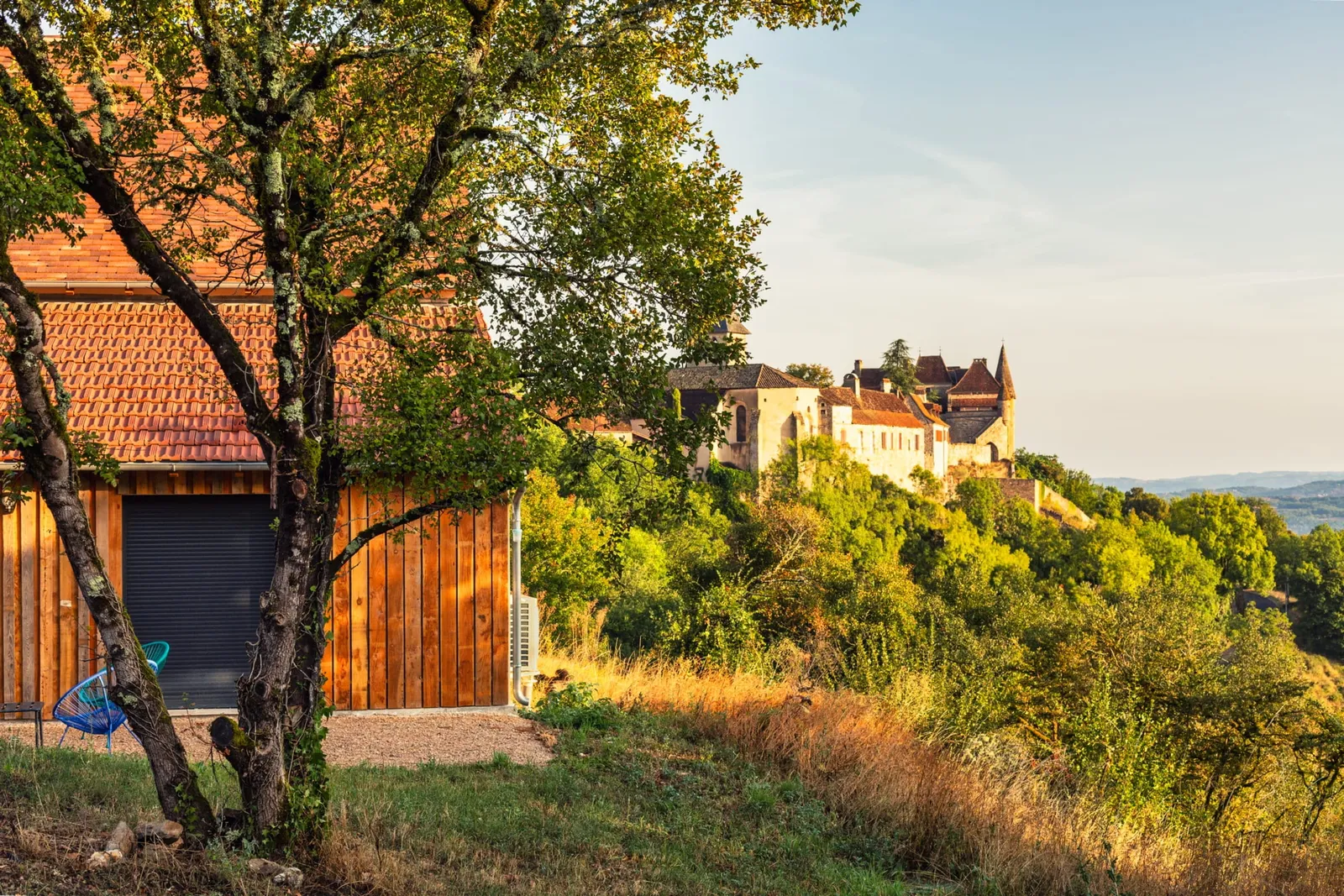 Grange au bardage en bois sous un arbre, avec le village de Loubressac en arrière-plan