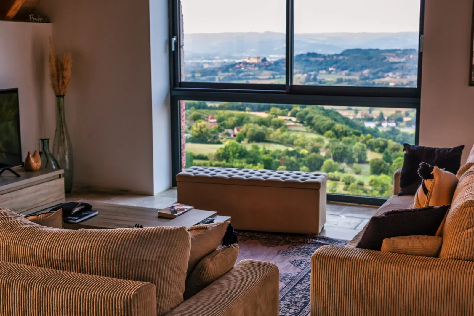 Salon aux canapés en velours côtelé devant une grande baie vitrée ouverte sur la vallée et un château au loin
