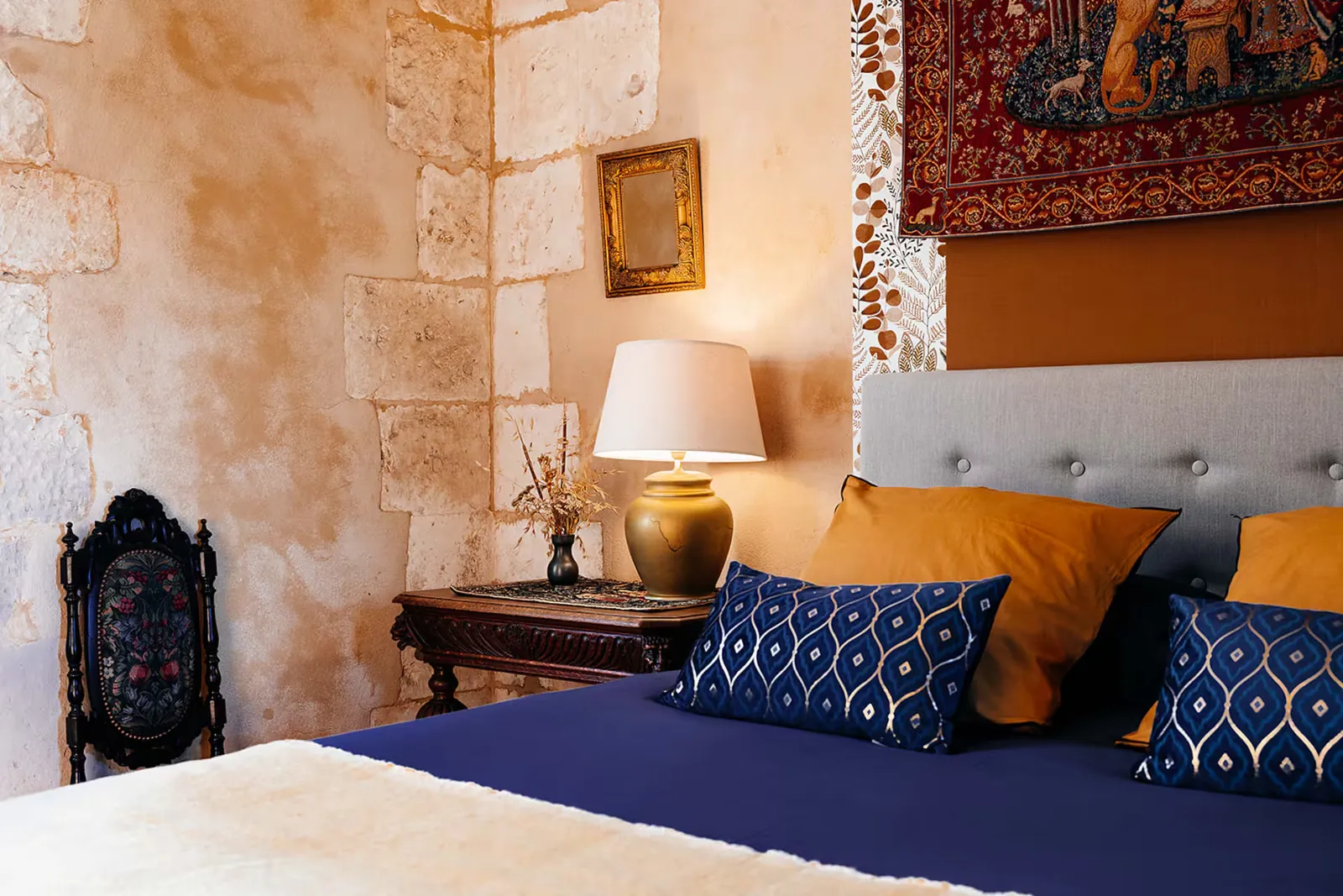 Chambre aux murs de pierre avec lit, coussins bleus et ocre et tapisserie