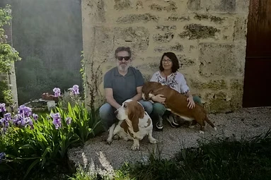 Eve, hôte de la Maison de la source, devant un mur en pierre fleuri d'iris, avec ses deux bassets