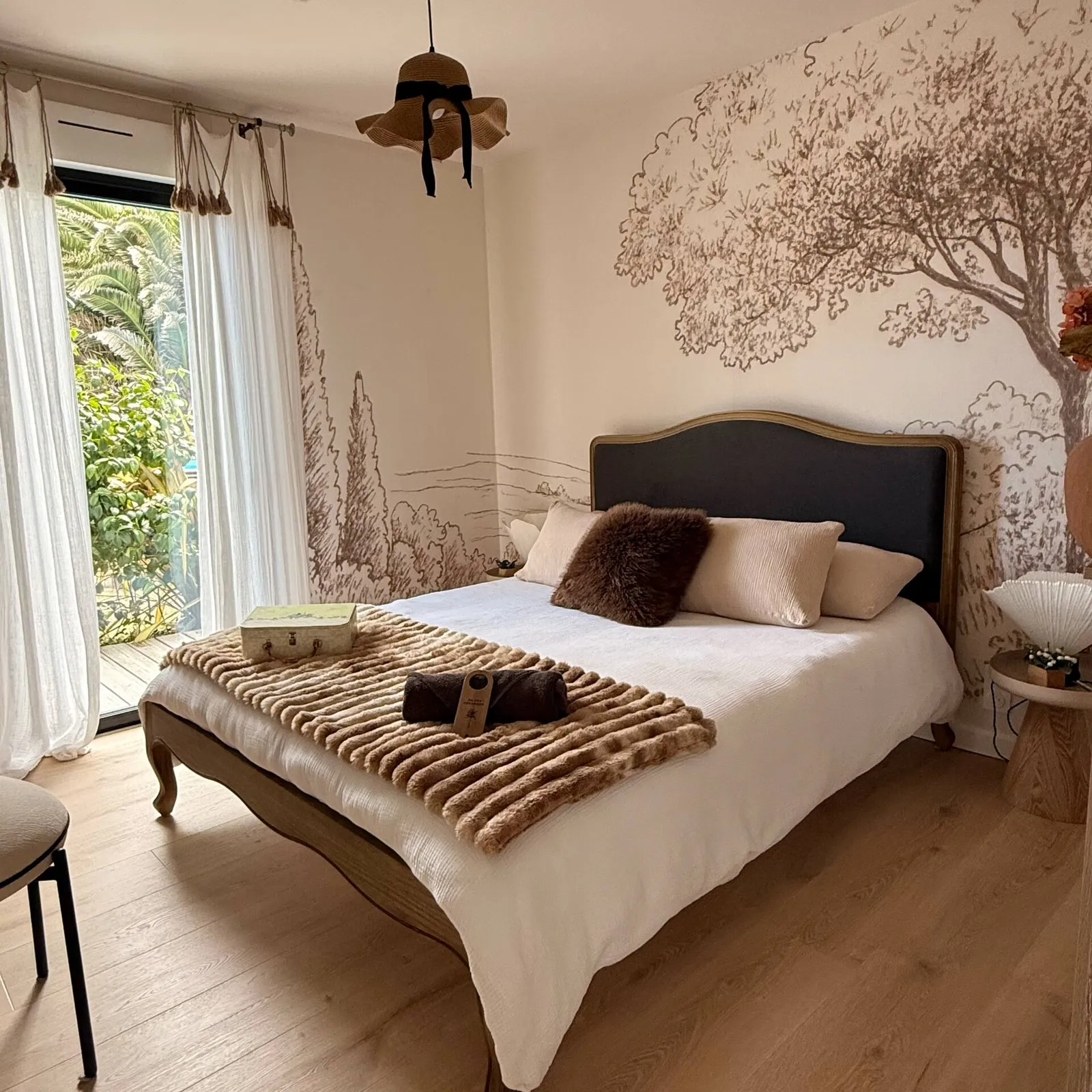 Chambre avec lit double, tête de lit capitonnée et fresque murale d'arbre, porte-fenêtre ouvrant sur le jardin