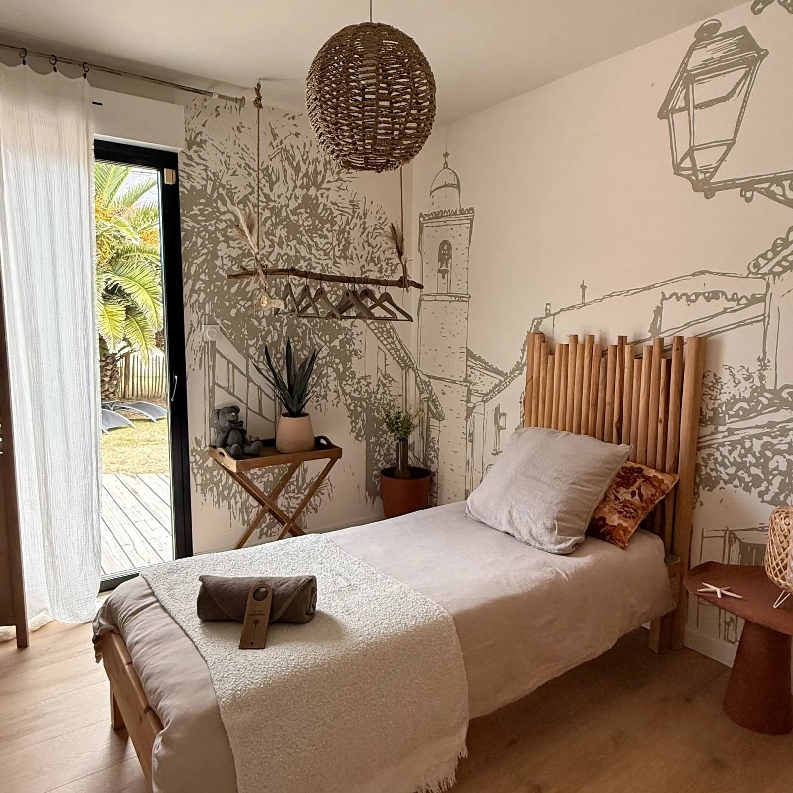 Chambre avec lit simple, tête de lit en rondins et fresque de village, porte-fenêtre ouvrant sur la terrasse