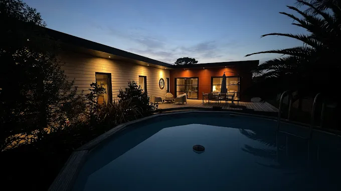 Façade éclairée de la Maison Goa à la tombée de la nuit, avec terrasse en bois et piscine au premier plan, à Portsall