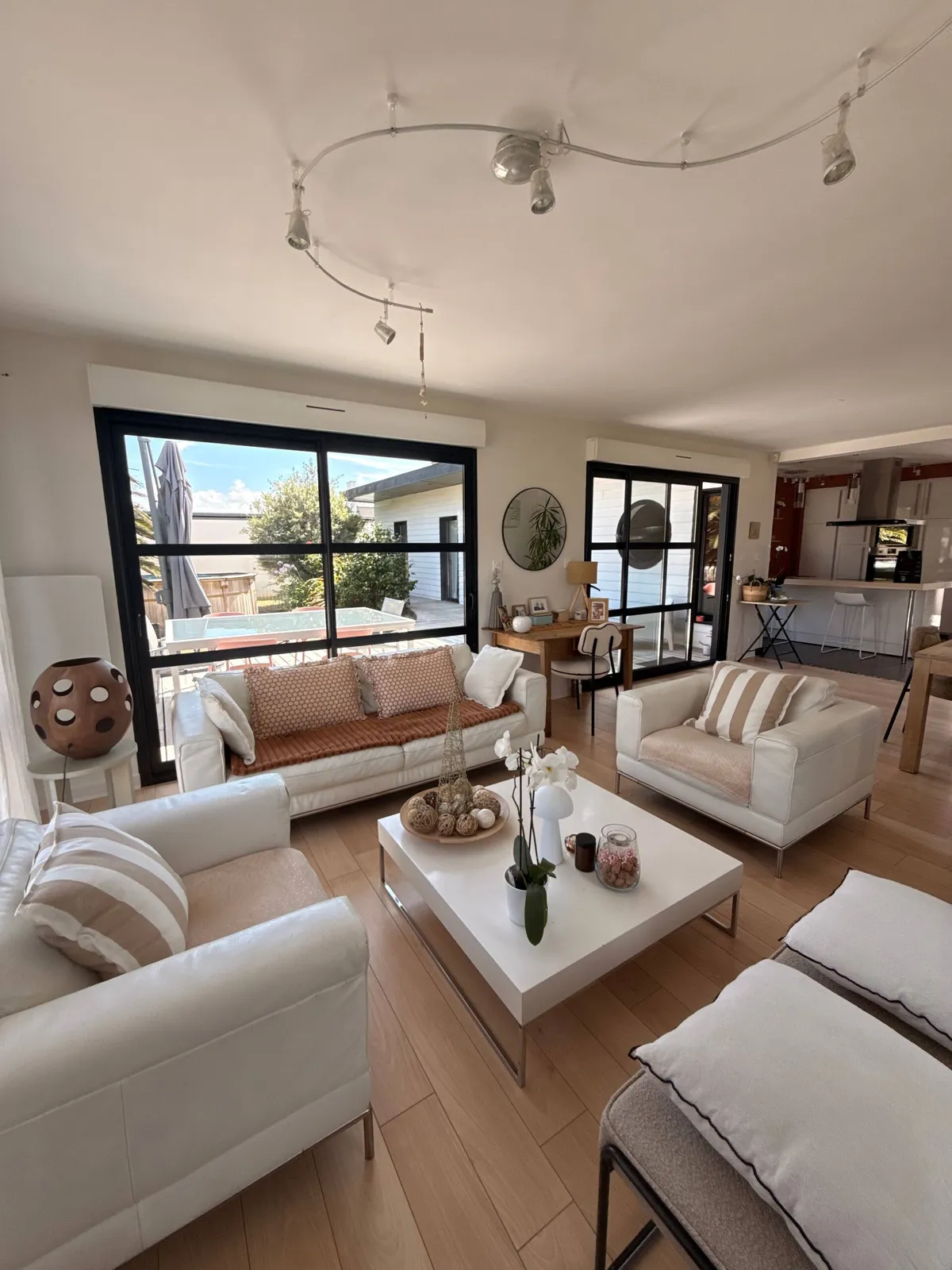 Séjour aux canapés blancs ouvert sur la terrasse par de grandes baies vitrées, avec la cuisine en arrière-plan